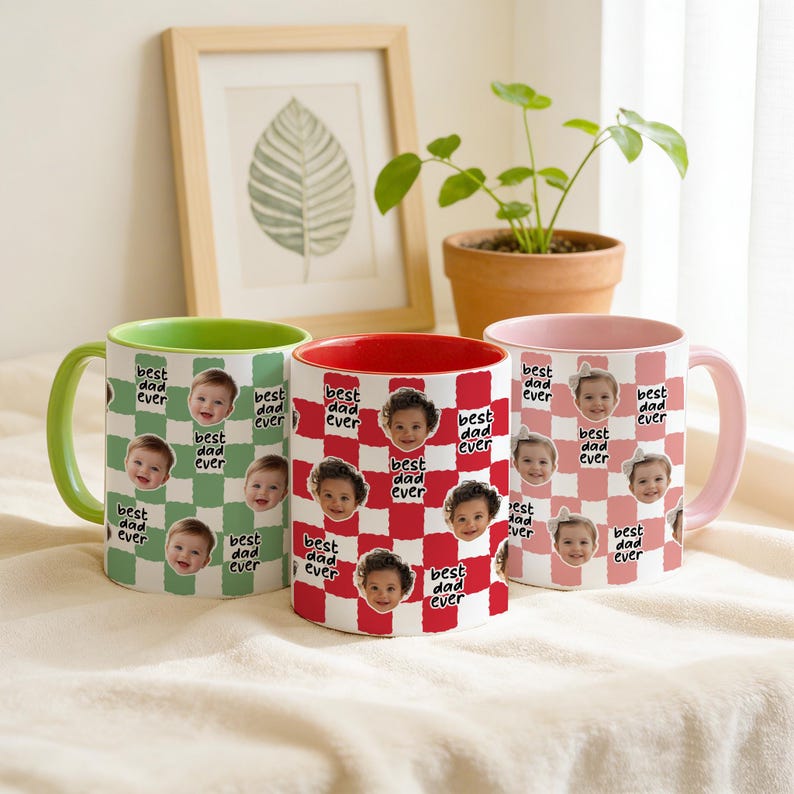 May include: Three ceramic mugs with the text "best dad ever" and baby faces on a checkered pattern. The mugs have green, red, and pink accents. A framed leaf print and a potted plant are in the background.