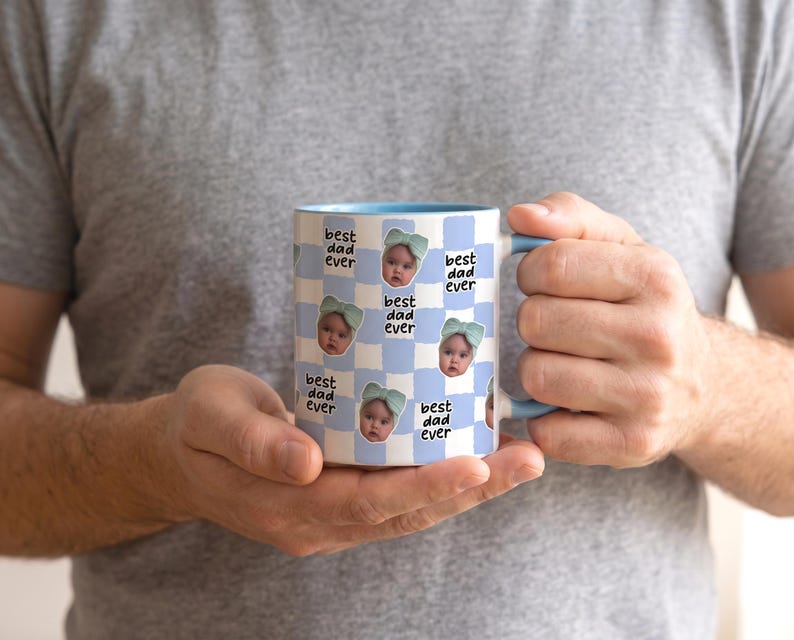 May include: A white ceramic mug with a light blue interior and handle. The mug features a repeating pattern of baby photos and the text "best dad ever" on a light blue checkered background. The mug is held by a person.