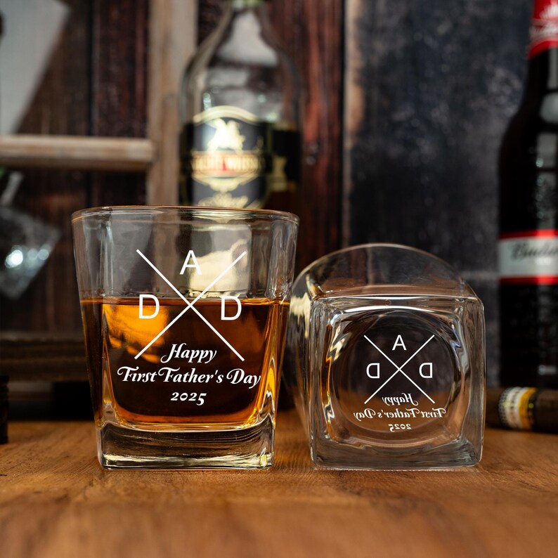 Op de afbeelding: Twee vierkante whiskyglazen gevuld met amberkleurige vloeistof. De glazen zijn ge&euml;tst met het woord "DAD" gekruist met een lijn en de tekst "Happy First Father's Day 2025". E&eacute;n glas staat rechtop, de andere ligt op zijn kant.