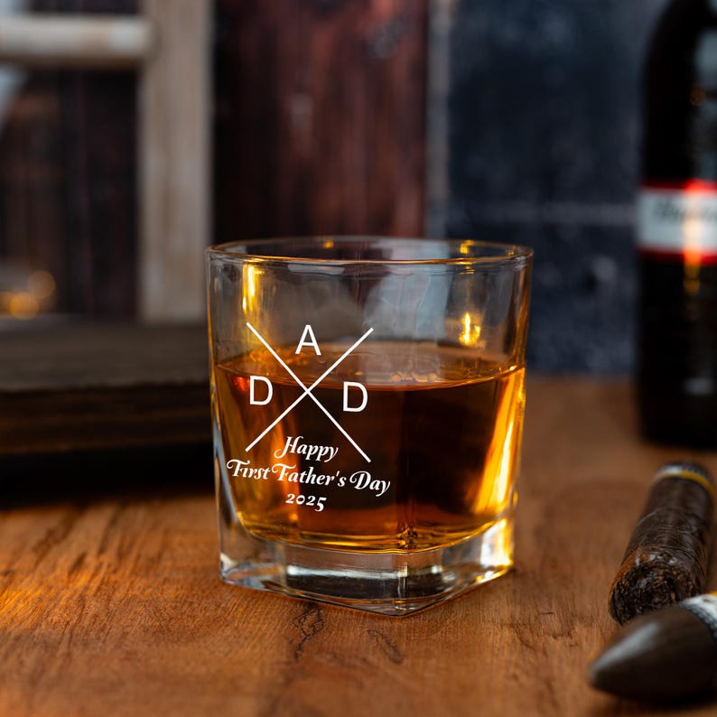 Op de afbeelding: Een vierkant glas gevuld met amberkleurige vloeistof, met de tekst "DAD" in een kruisontwerp en "Happy First Father's Day 2025". Het glas staat op een houten oppervlak, met een sigaar en een fles op de achtergrond.