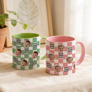 May include: Two ceramic mugs with a checkered pattern and the text "best dad ever". One mug is green and white with a green handle, and the other is pink and white with a pink handle. Each mug features a baby's face.