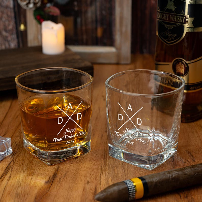 Op de afbeelding: Twee vierkante whiskyglazen, &eacute;&eacute;n gevuld met amberkleurige vloeistof, de andere leeg. Beide glazen zijn ge&euml;tst met een ontwerp met de gekruiste letters "A" en "D", en de tekst "Happy First Father's Day 2025". Een sigaar en een fles whisky staan op de achtergrond.