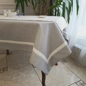 May include: A gray linen tablecloth with a white border and trim. The tablecloth is draped over a wooden table. A white runner is on top of the tablecloth. A small white dish and a decorative vase are on the table.