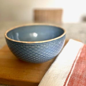 May include: A round ceramic bowl with a dusty blue glaze and a textured, scale-like pattern on the exterior. The rim of the bowl features a contrasting light beige or tan edge, highlighting the depth of the blue color and the detailed surface.