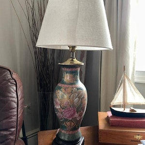 Può includere: Una lampada da tavolo con paralume conico in tessuto bianco e base in ceramica su fondo verde scuro, decorata con un motivo floreale colorato nei toni del rosa, arancione e oro, poggiante su una base quadrata in legno marrone scuro.