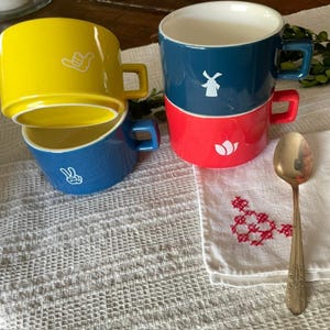 May include: Stack of three ceramic mugs in yellow, blue, and red. The yellow mug has a white hand gesture, the blue mug has a white peace sign, and the red mug has a white tulip. A gold spoon rests on a white cloth.