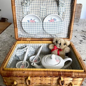 May include: A woven wicker picnic basket with a blue and white checkered fabric lining. Inside are two small white ceramic plates, two white ceramic spoons, a white teapot with floral accents, and a small beige teddy bear with a red ribbon.