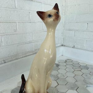 May include: A tall, slender ceramic Siamese cat figurine with a cream-colored body, dark brown ears, face, paws, and tail. The cat has blue eyes, long painted eyelashes, and is depicted in a seated position with its head tilted upward.