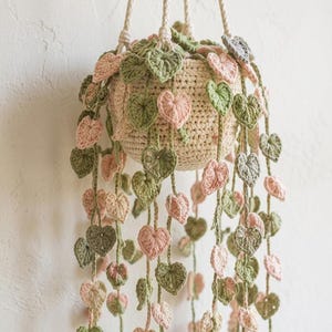 May include: A crocheted hanging plant featuring a cream-colored pot and long, trailing vines adorned with numerous small, heart-shaped leaves in alternating shades of soft sage green and pale pastel pink, suspended by thick, braided cream-colored cords.