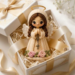 May include: A crocheted angel doll with brown braided hair, a cream-colored hooded robe, and white wings. The doll features a gold halo, a small black rosary with a cross, and pink crocheted roses at the hem, presented inside a white gift box.