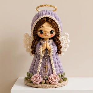 May include: A crocheted angel doll with brown braided hair, wearing a lavender hooded robe with white trim. The figure has white wings, a gold halo, and holds a rosary with a cross. The base is decorated with three pink roses and green leaves.
