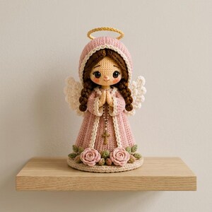 May include: A crocheted angel doll with brown hair in braids, wearing a pink hooded robe with white trim. The doll has white wings, a gold halo, and holds a small rosary with a cross. The base is decorated with pink roses and green leaves.