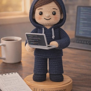 May include: A crocheted doll of a person in a navy blue hoodie and trousers, holding a small laptop displaying code. The doll stands on a wooden base with the text "IT / Programmer". A coffee mug and notebook are in the background.
