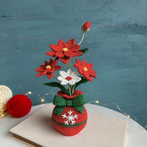 May include: A crocheted poinsettia arrangement in a red vase with a green bow and snowflake detail. The flowers are red with yellow centres and white accents. The arrangement sits on a neutral surface against a teal background.