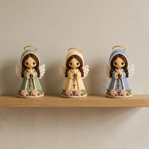 May include: Three crocheted angel figurines with brown hair, gold halos, and white wings. Each figure wears a long dress in sage green, cream, or light blue, featuring a rosary with a cross, and is decorated with pink roses at the base.