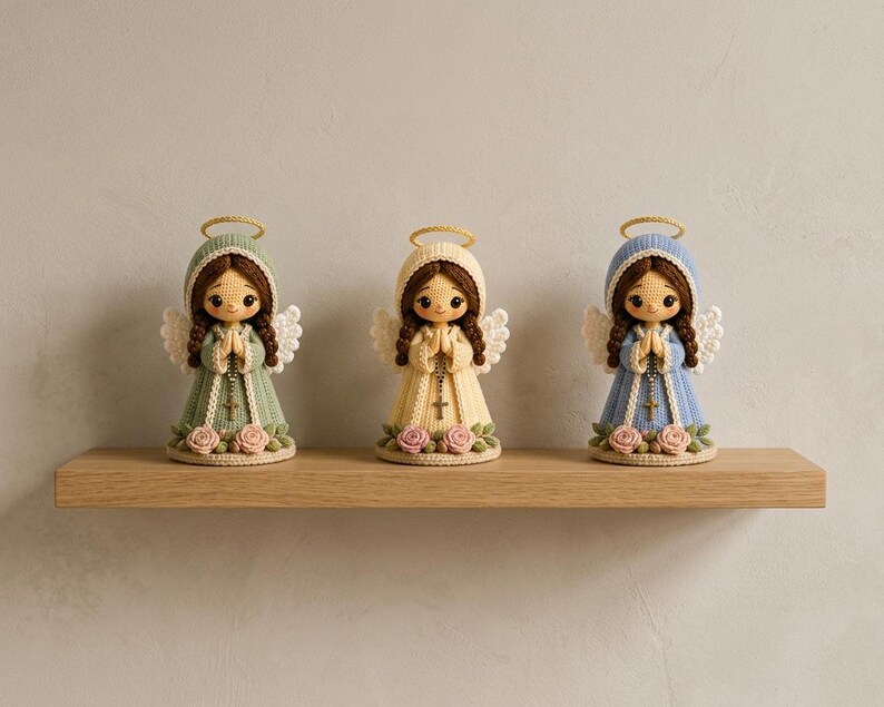May include: Three crocheted angel figurines with brown hair, gold halos, and white wings. Each figure wears a long dress in sage green, cream, or light blue, featuring a rosary with a cross, and is decorated with pink roses at the base.