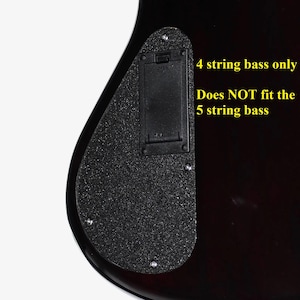 May include: A black bass guitar battery compartment cover with a textured finish, secured by four screws. The text on the right reads: 4 string bass only. Does NOT fit the 5 string bass. The component is mounted on a dark, glossy instrument body.
