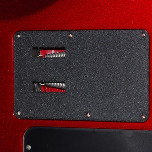 May include: Close-up of a black rectangular plate with two spring-loaded access panels. The plate is mounted on a red surface.