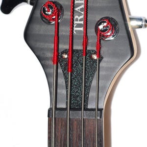 May include: The headstock of a bass guitar featuring a matte black finish with a textured black plate. Four silver metal strings are wound around chrome tuning pegs, each accented with bright red silk winding at the top and near the nut.