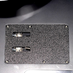 May include: A rectangular, textured black metal plate with two recessed compartments, each containing a metal component with a spring and screw. The plate has six small, evenly spaced holes around the edges.