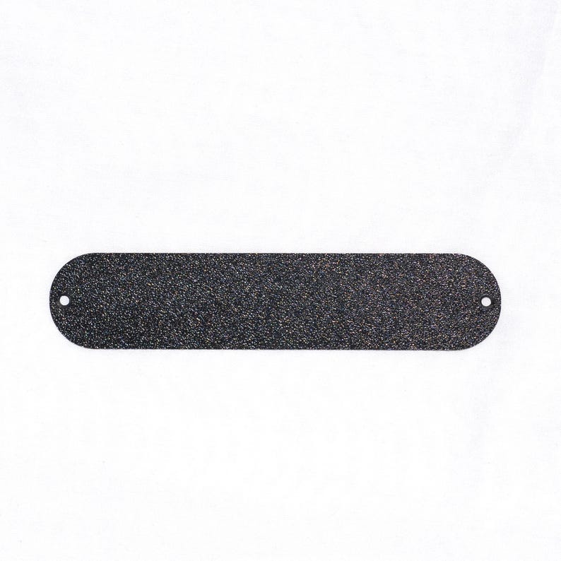 May include: A long, oval-shaped, black metal plate with a textured surface and rounded ends. Two small holes are drilled near the ends. The plate is set against a white background.