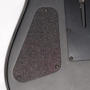 May include: Close-up of a black guitar body with a textured, dark gray backplate secured by four screws. The guitar's edge is visible, showcasing its sleek, dark finish. The backplate has a speckled appearance.
