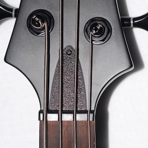 May include: Close-up of a black bass guitar headstock. The headstock has a unique shape with tuning pegs and strings visible. The fretboard is a dark brown color, and the strings are silver.