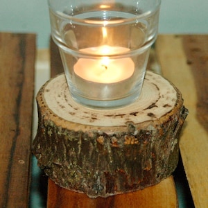 May include: A small, round, wooden candle holder with a natural bark finish. A glass votive candle holder with a lit tea light sits on top of the wooden holder.