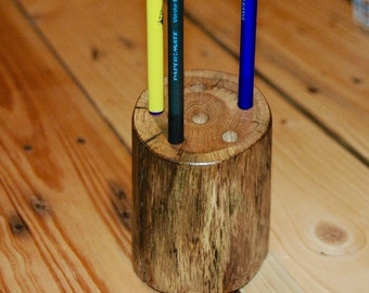 May include: A wooden pencil holder with three holes, made from a tree trunk. The holder has a dark brown stain and is holding a yellow, blue, and black pen.