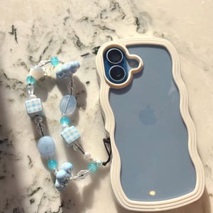 May include: A light blue phone case with a white wavy border and a matching beaded phone charm. The charm features blue beads, checkered squares, and cloud-shaped accents. The phone case has a clear back.