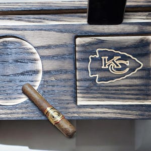 May include: A wooden cigar and phone stand with a circular ashtray, a phone holder, and a rectangular area with the text "KC" in a stylized arrowhead design.