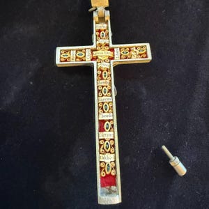 May include: A silver-toned metal cross pendant featuring a red velvet interior inlay. The cross is decorated with intricate gold-colored filigree scrollwork, small green accents, and paper labels with text including Agnus Dei, Marcus, and Theodo.