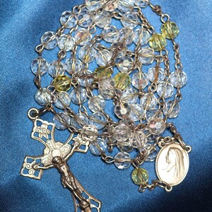 May include: A vintage rosary with clear, faceted beads and silver-toned metal chain. The crucifix features a detailed depiction of Jesus, and the oval pendant shows a profile of the Virgin Mary. Some beads are yellow.