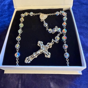 May include: A rosary with iridescent crystal beads and a silver crucifix, presented in a white box with a black interior. The crucifix features a detailed depiction of Jesus. The beads are connected by a delicate silver chain.