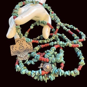 May include: A long necklace featuring strands of irregular turquoise chips and small red beads. The necklace includes a large, rectangular silver-toned metal cross pendant with an embossed heart and cross design, plus a circular silver-toned metal charm.