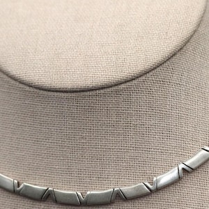 May include: A silver-toned metal necklace featuring a series of repeating, angled geometric links. Each link has a smooth, polished finish with a distinct V-shaped notch, creating a continuous, segmented chain design that rests in a curved shape.