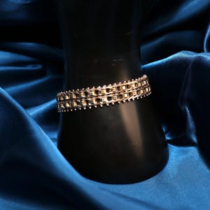 May include: A gold-colored bracelet with a chain-like design. The bracelet is composed of small, interconnected rectangular links, creating a textured surface. The jewelry is displayed on a black form against a backdrop of blue fabric.
