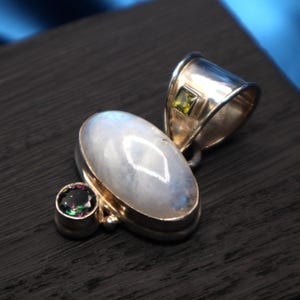 May include: A silver pendant featuring a large, oval moonstone with a diamond-shaped inclusion. A smaller, round, iridescent gemstone and a square, green gemstone are also set in the silver. The pendant has a polished finish.