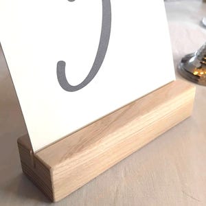 Peut inclure: Un support rectangulaire en bois clair avec une fente horizontale sur le dessus, maintenant une carte blanche sur laquelle est imprimé le chiffre 3 en gris foncé avec une police cursive. La base en bois présente un grain naturel visible.