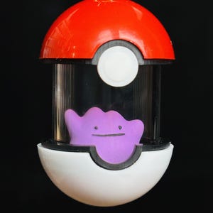 May include: A red and white Poké Ball-shaped pendant with a gold-colored loop at the top. Inside the clear cylindrical section is a purple Ditto figure. The pendant is a collectible item.