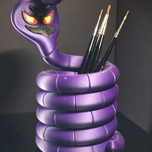 Puede incluir: Un organizador de escritorio morado con forma de serpiente, con un cuerpo enrollado y una cabeza de cobra. La cabeza tiene ojos rojos brillantes y contiene pinceles. El organizador está hecho de un material liso y sólido.
