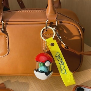 May include: A red, white, and black Poké Ball keyring with a Bulbasaur figure inside. The keyring has a yellow strap with the word "Pokémon" in green. The background features a brown leather handbag.