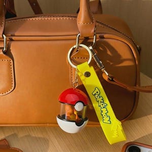 May include: A red, white, and black Pokemon-themed keychain with a yellow strap that says "Pokemon". The keychain is attached to a brown leather handbag.