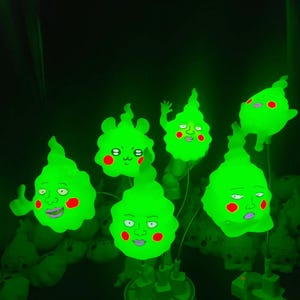 Op de afbeelding: Een cluster van gloeiende groene cartoonkarakterlampen met expressieve gezichten en rode wangen. De lampen zijn in een groep gerangschikt en geven een heldergroen licht af tegen een donkere achtergrond. De lampen hebben een uniek, grillig ontwerp.