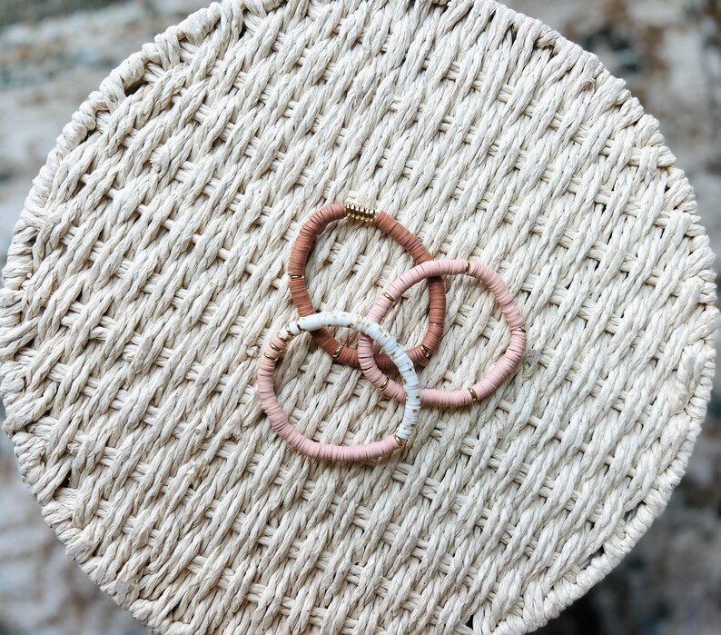 May include: Three bracelets made of small, round beads in shades of pink and white. The bracelets are stacked on top of each other and have a gold accent.
