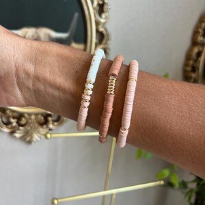 May include: Three bracelets made of small, round, colourful beads. The bracelets are stacked on a person's wrist. The beads are white, light pink, and a darker pink.