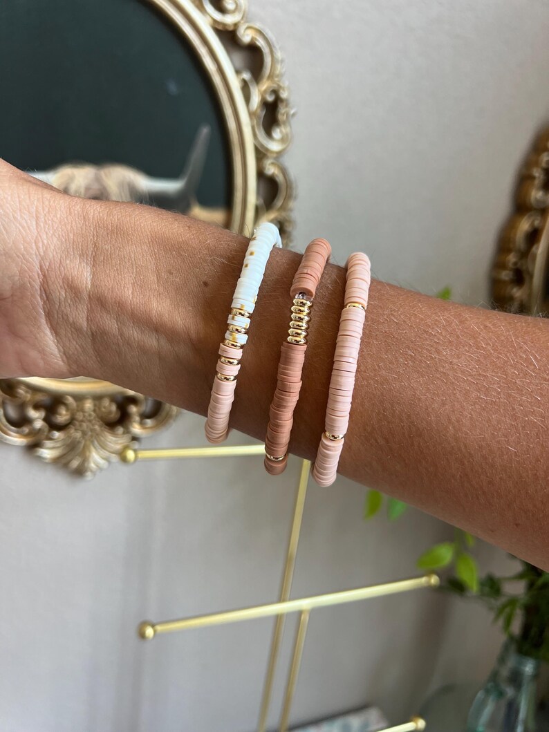 May include: Three bracelets made of small, round, colourful beads. The bracelets are stacked on a person's wrist. The beads are white, light pink, and a darker pink.