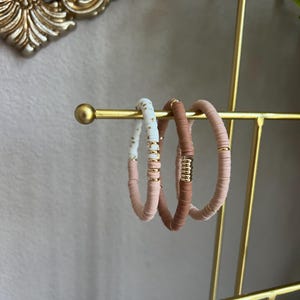 May include: Three bracelets on a gold stand. The bracelets are made of small, round beads in shades of pink, white, and gold.