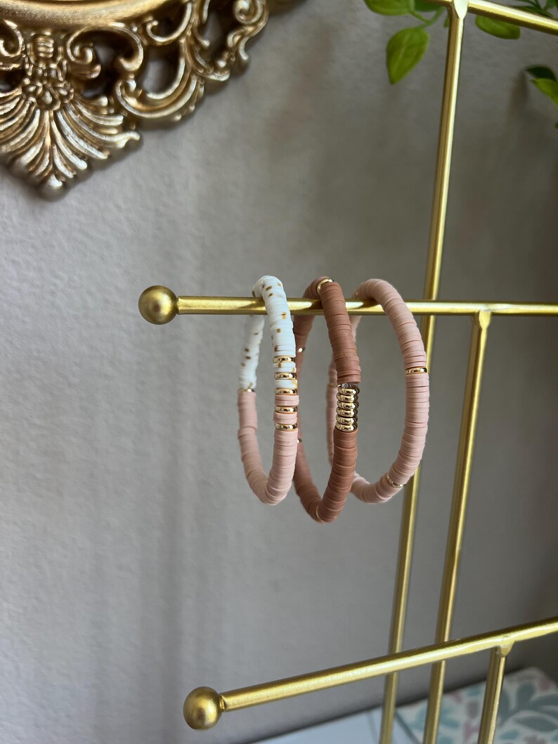 May include: Three bracelets on a gold stand. The bracelets are made of small, round beads in shades of pink, white, and gold.