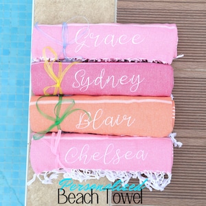 Puede incluir: Cuatro toallas de playa rosas y naranjas están apiladas sobre una superficie de madera. Cada toalla está personalizada con un nombre: Grace, Sydney, Blair y Chelsea. Las toallas tienen flecos blancos y están atadas con cintas de colores. El texto "Toalla de playa personalizada" es visible en la parte inferior de la imagen.